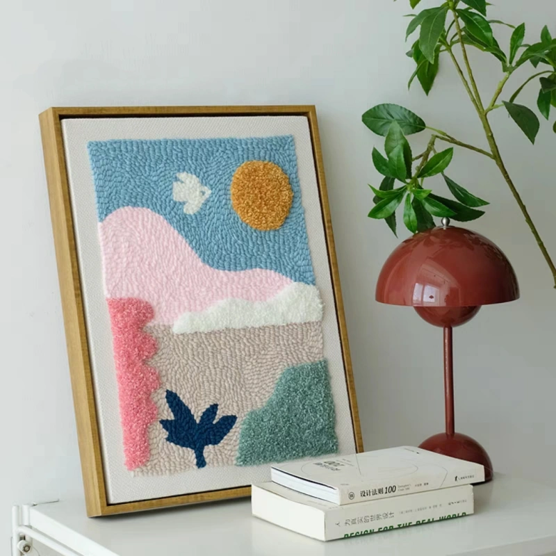 Sunset Valley Punch Needle Wall Art Kit – Abstract Decor Framed punch needle wall art featuring an abstract sunset valley scene with pink hills, blue sky, and sun, displayed on a shelf.