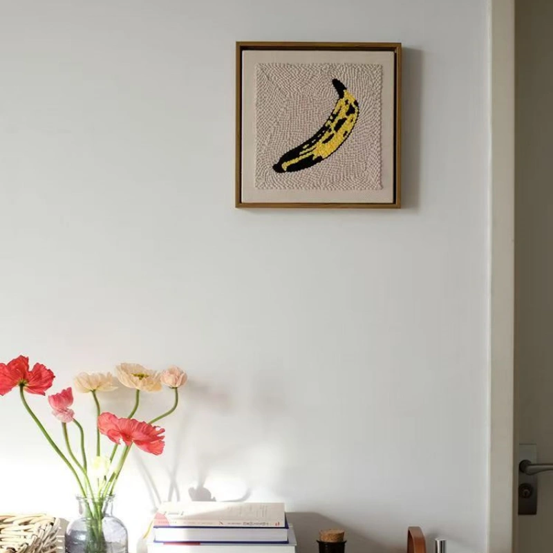 Framed Andy Warhol Banana Punch Needle Art - Modern Wall Decor Framed Andy Warhol banana punch needle artwork hanging on white wall above flowers and books, modern interior design