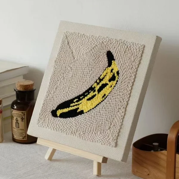 Andy Warhol Banana Punch Needle Wall Art Kit - Pop Art DIY Canvas Andy Warhol inspired yellow banana punch needle art on beige canvas displayed on wooden easel, pop art home decor