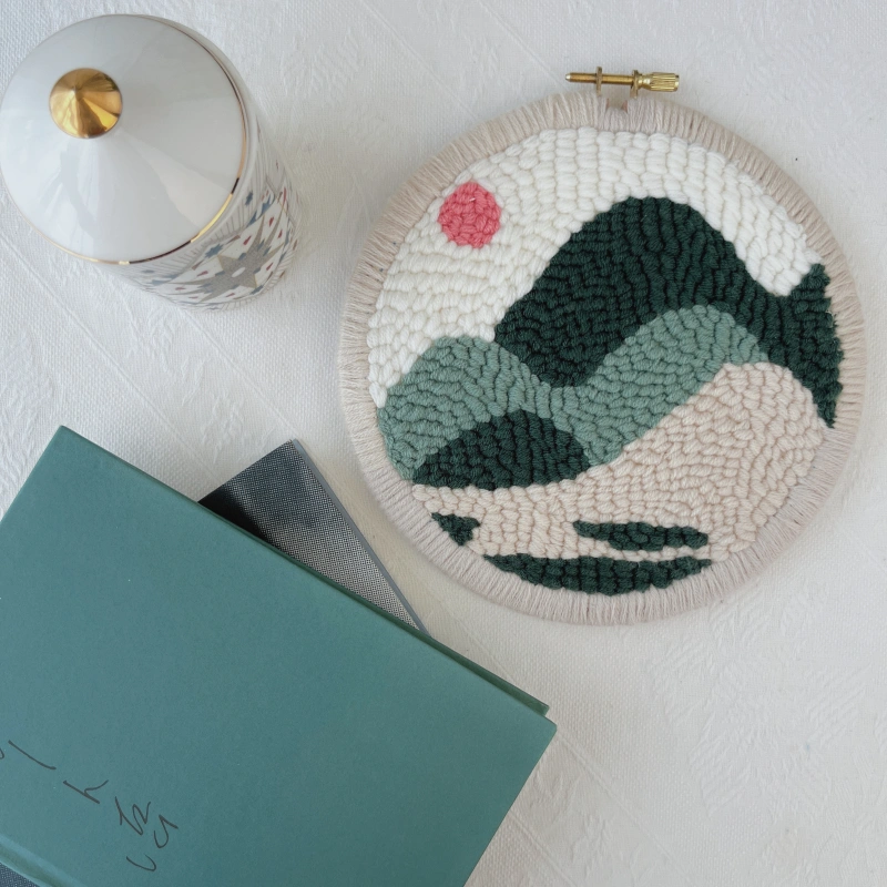 Finished hoop wall art with edge binding, showing the Holakits Mountain Landscape design displayed beside two hardcover books and a scented candle.