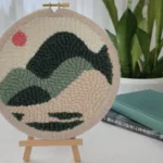 Finished hoop wall art with edge binding displayed on a wooden tripod stand as a desktop décor piece, with books and a plant in the background.