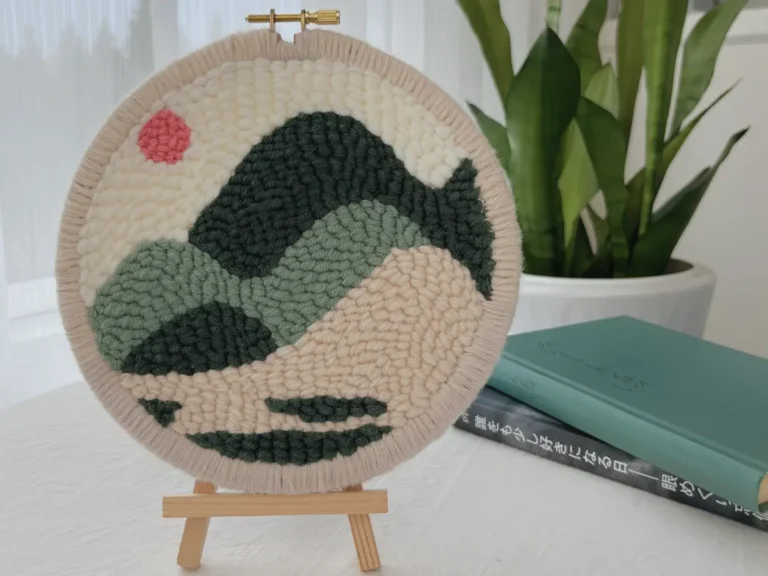 Finished hoop wall art with edge binding displayed on a wooden tripod stand as a desktop décor piece, with books and a plant in the background.