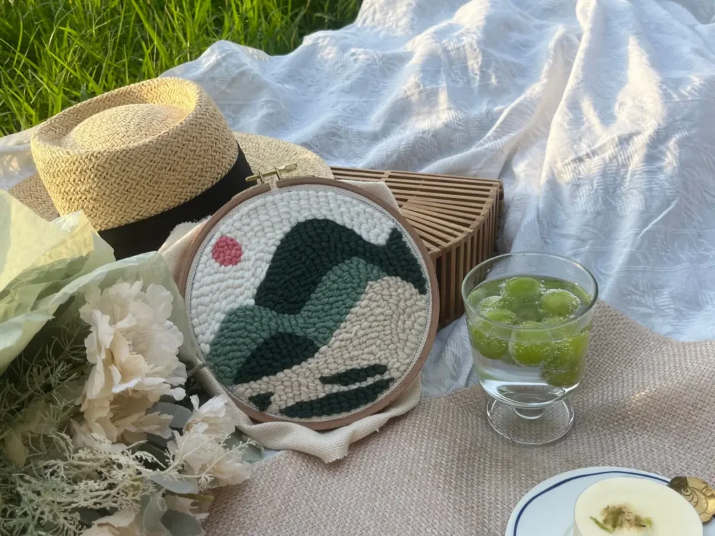 Finished hoop artwork without edge binding placed casually on a picnic blanket on grass, surrounded by flowers, a straw hat, sparkling grape drink, and a woven bag.