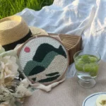Finished hoop artwork without edge binding placed casually on a picnic blanket on grass, surrounded by flowers, a straw hat, sparkling grape drink, and a woven bag.