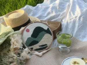 Finished hoop artwork without edge binding placed casually on a picnic blanket on grass, surrounded by flowers, a straw hat, sparkling grape drink, and a woven bag.