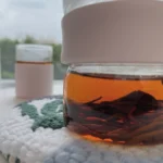 Close-up of a finished Holakits Wild Leaves hoop trivet holding a teapot of red tea, highlighting the thick texture, with a pink cup and bright window view in the background.