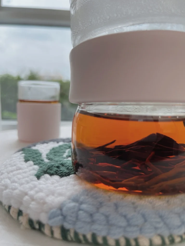 Close-up of a finished Holakits Wild Leaves hoop trivet holding a teapot of red tea, highlighting the thick texture, with a pink cup and bright window view in the background.
